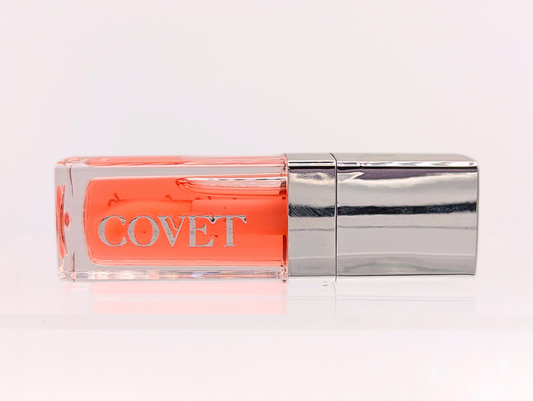 ‘Want’ lip gloss by Covet Beauty, a warm coral shade with a high mirror shine. The gloss lies horizontally on a soft white background with a silver cap and subtle reflection below.