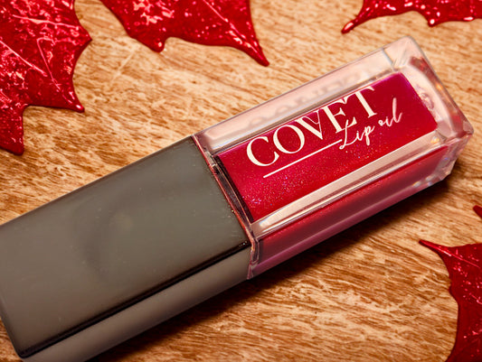 Covet Maple Love lip gloss styled on a wood surface with red maple leaves, showcasing a warm crimson gloss in a clear rectangular tube.