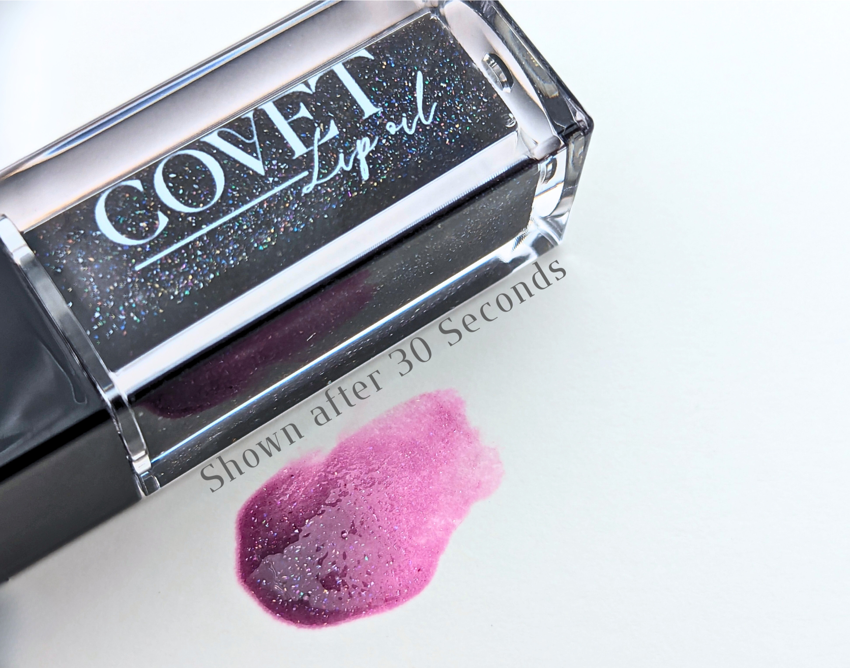 Covet Beauty Black Currant lip gloss — a deep black-violet gloss with subtle iridescent shimmer that blooms into a cool berry flush, shown swatched after 30 seconds on a white background