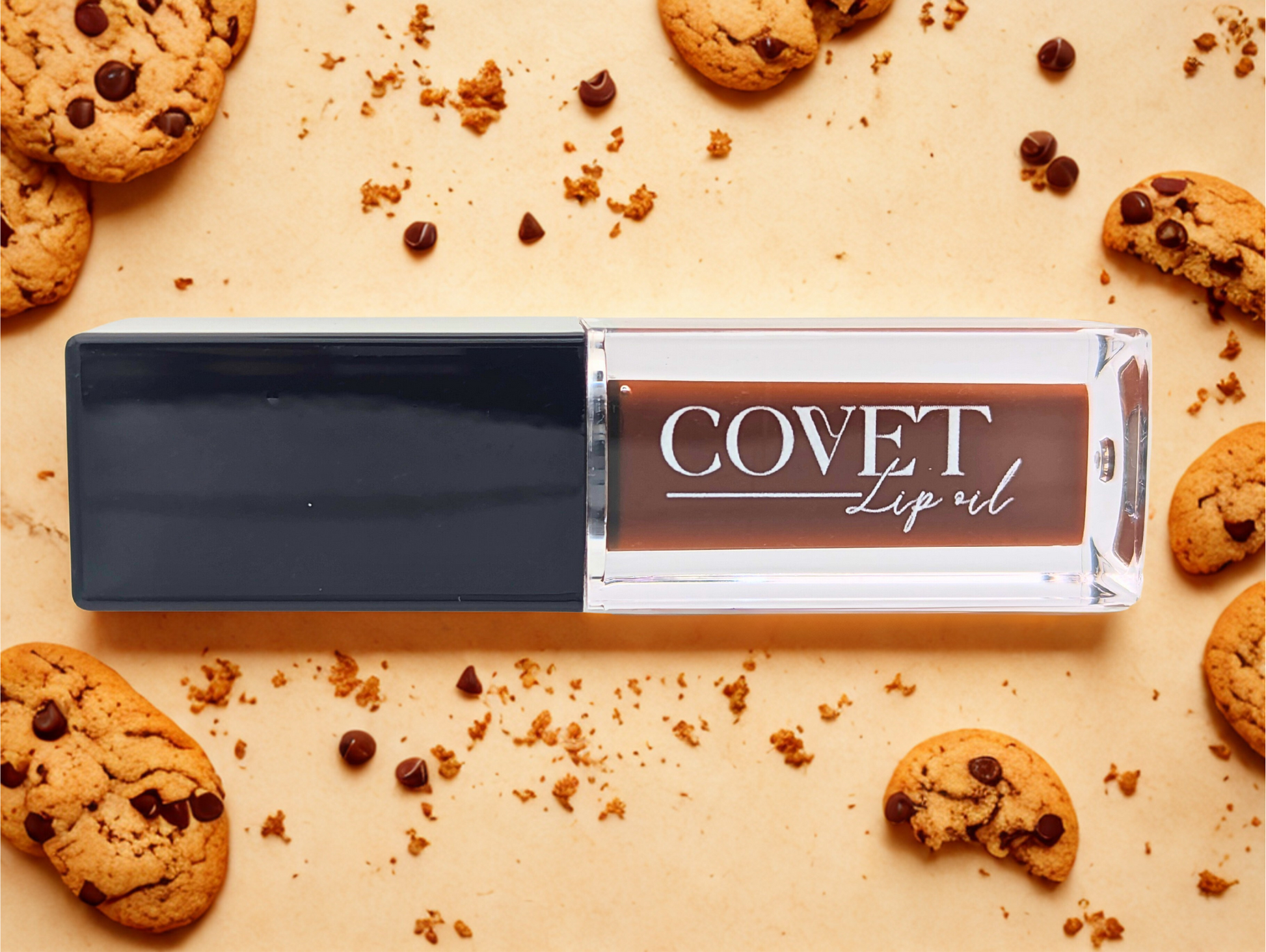 Covet Beauty’s Chocolate Cookie lip gloss — a creamy medium brown gloss in a clear rectangular tube with black cap, shown on a cookie-themed background.