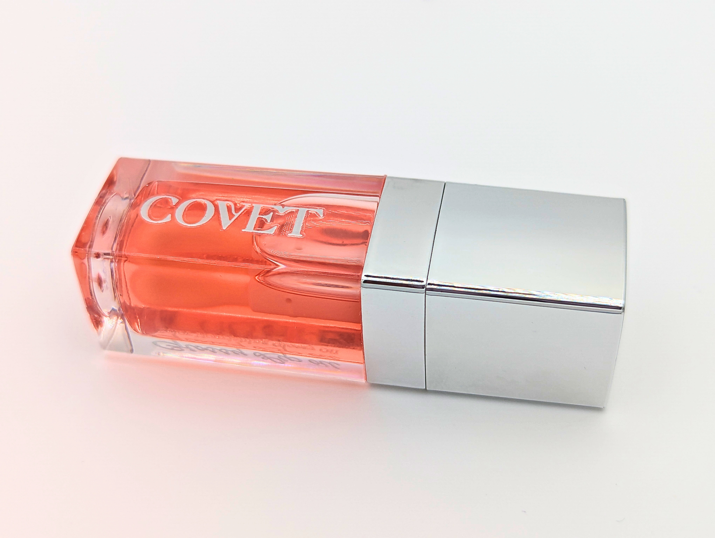 Close-up angled view of ‘Want’ lip gloss by Covet Beauty, showing its warm coral tone and glassy, high-shine finish. The tube rests on a white surface with soft lighting and a silver cap reflection.