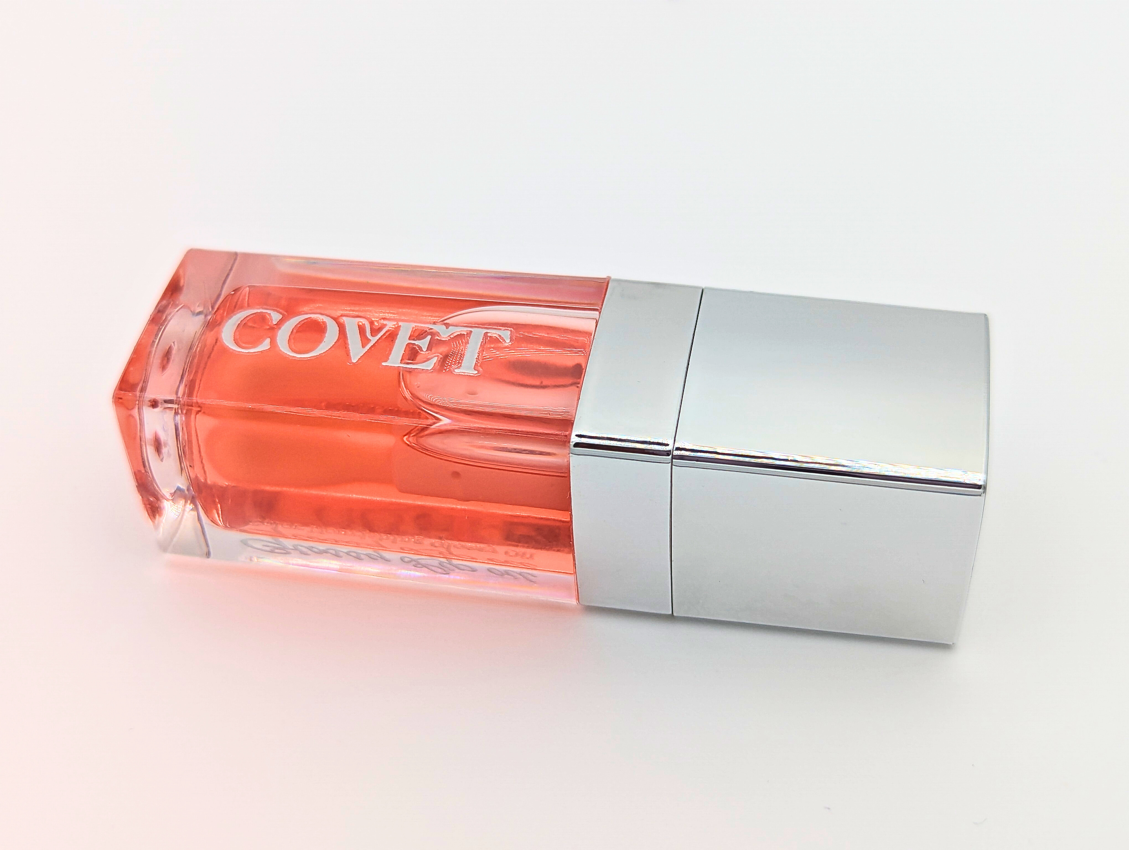 Close-up angled view of ‘Want’ lip gloss by Covet Beauty, showing its warm coral tone and glassy, high-shine finish. The tube rests on a white surface with soft lighting and a silver cap reflection.
