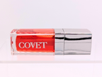 ‘Yearn’ lip gloss by Covet Beauty, a fiery coral-red shade with warm undertones and a radiant mirror finish. The gloss lies horizontally on a white background with a silver cap and soft reflection below.