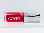 ‘Crave’ lip gloss by Covet Beauty, a cool-toned raspberry shade with a high mirror finish. The gloss rests horizontally on a clean white background with a silver cap and subtle reflection beneath.
