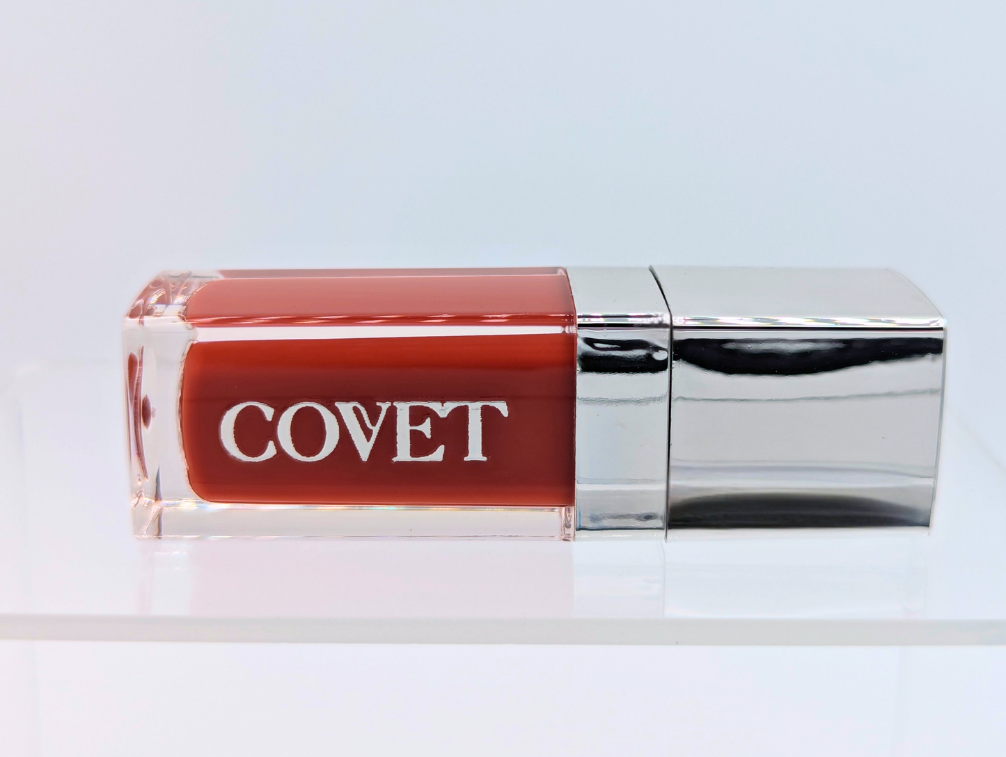 ‘Melt’ lip gloss by Covet Beauty, a rich terracotta shade with golden undertones and a high mirror finish. The gloss lies horizontally on a white background with a silver cap and soft reflection beneath.