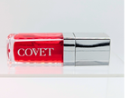 ‘Fever’ lip gloss by Covet Beauty, a true red shade with warm undertones and a high mirror finish. The gloss lies horizontally on a clean white background with a silver cap and soft reflection beneath.