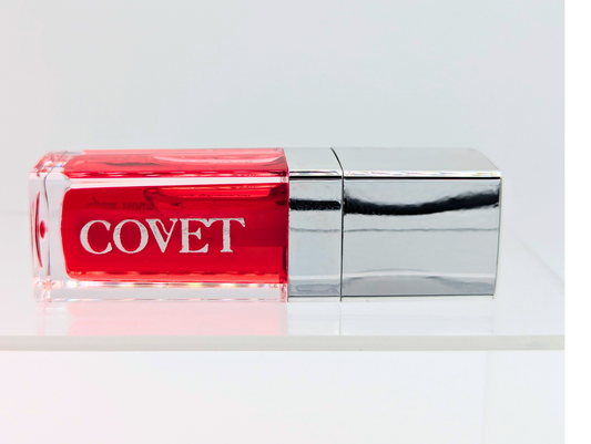 ‘Fever’ lip gloss by Covet Beauty, a true red shade with warm undertones and a high mirror finish. The gloss lies horizontally on a clean white background with a silver cap and soft reflection beneath.