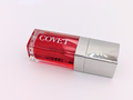 Angled view of ‘Fever’ lip gloss by Covet Beauty, showing its warm red tone and glossy, reflective finish. The clear tube catches soft light on a white surface, highlighting its vivid color and smooth texture.