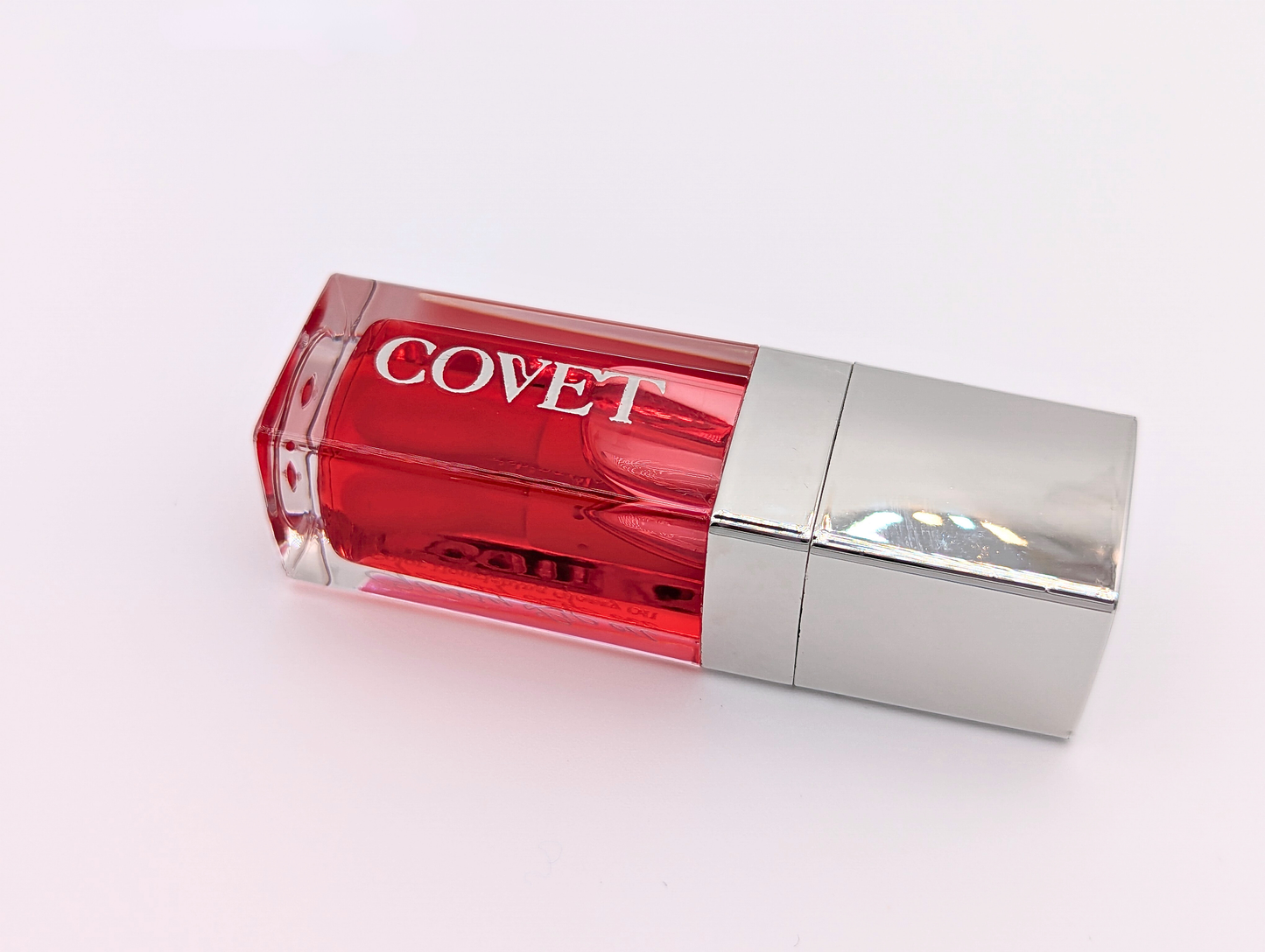 Angled view of ‘Fever’ lip gloss by Covet Beauty, showing its warm red tone and glossy, reflective finish. The clear tube catches soft light on a white surface, highlighting its vivid color and smooth texture.
