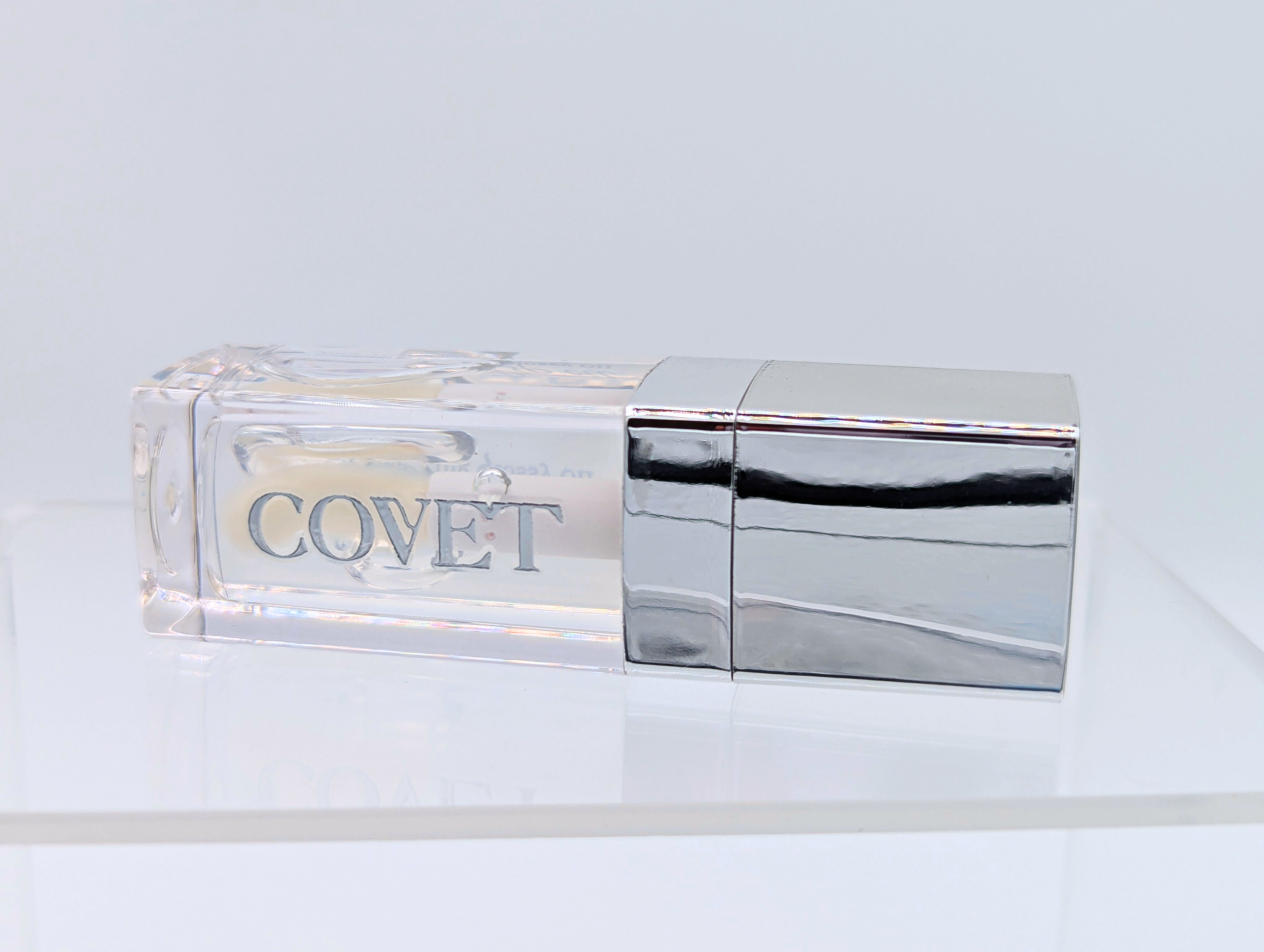 Angled view of ‘Touch’ lip gloss by Covet Beauty, showing its clear, mirror-like finish and sleek reflective tube. The gloss rests on a white surface under soft light, highlighting its smooth, glassy texture.