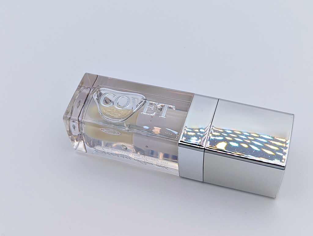 Angled view of ‘Touch’ lip gloss by Covet Beauty, showing its clear, mirror-like finish and sleek reflective tube. The gloss rests on a white surface under soft light, highlighting its smooth, glassy texture.