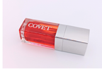Angled view of ‘Yearn’ lip gloss by Covet Beauty, showing its warm coral-red tone and glossy, high-shine finish. The clear tube reflects soft light on a white surface, highlighting its smooth, luxurious texture.