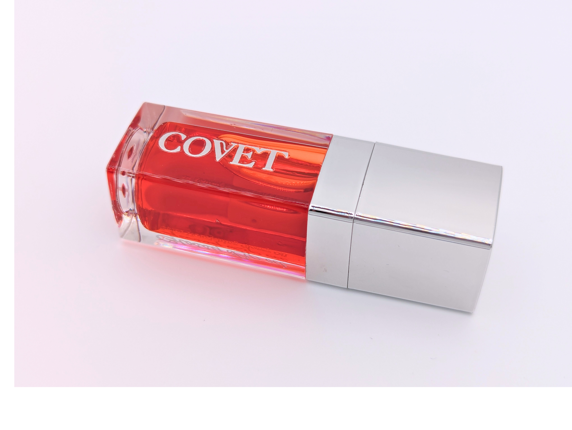 Angled view of ‘Yearn’ lip gloss by Covet Beauty, showing its warm coral-red tone and glossy, high-shine finish. The clear tube reflects soft light on a white surface, highlighting its smooth, luxurious texture.