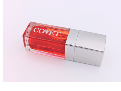 Angled view of ‘Yearn’ lip gloss by Covet Beauty, showing its warm coral-red tone and glossy, high-shine finish. The clear tube reflects soft light on a white surface, highlighting its smooth, luxurious texture.