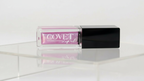 A Covet Lip Oil in a clear rectangular tube with a black cap, filled with a shimmering violet-pink shade, photographed on a glossy white surface with soft, diffused lighting.