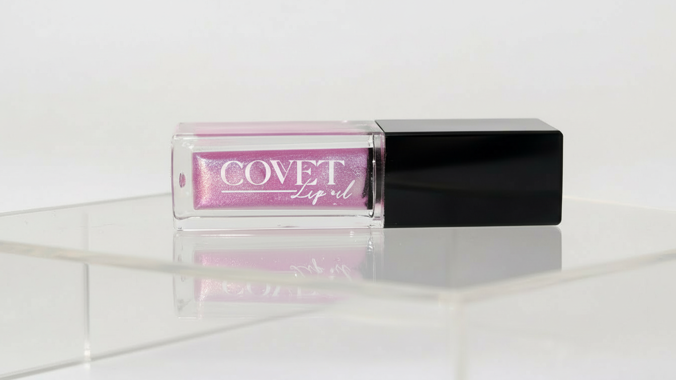 A Covet Lip Oil in a clear rectangular tube with a black cap, filled with a shimmering violet-pink shade, photographed on a glossy white surface with soft, diffused lighting.