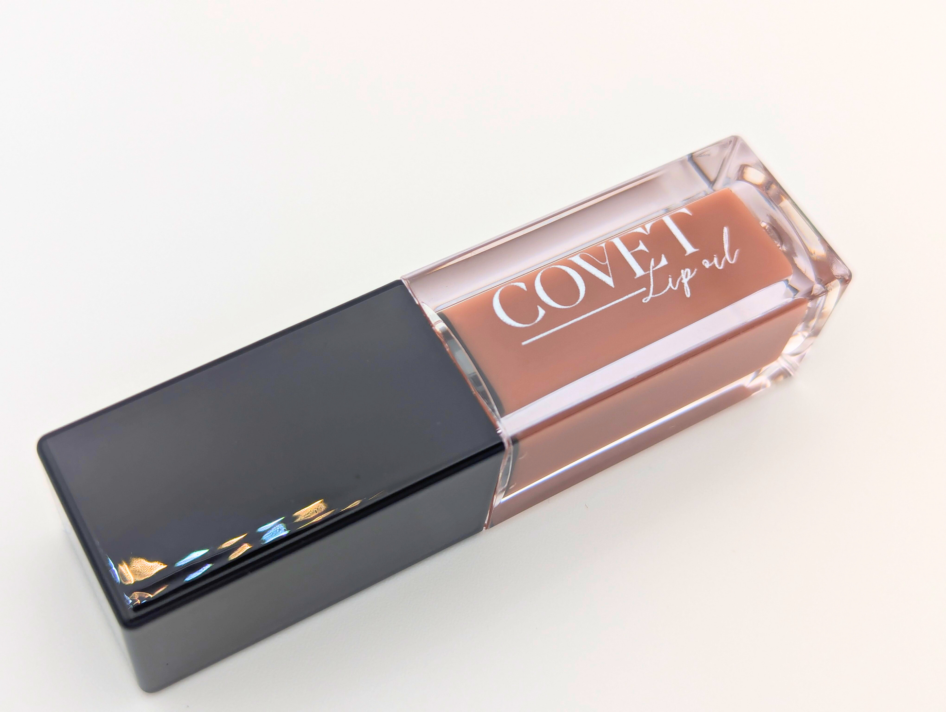 Angled view of Covet Latte Cream lip gloss on a white background, displaying the warm nude tone in a transparent tube