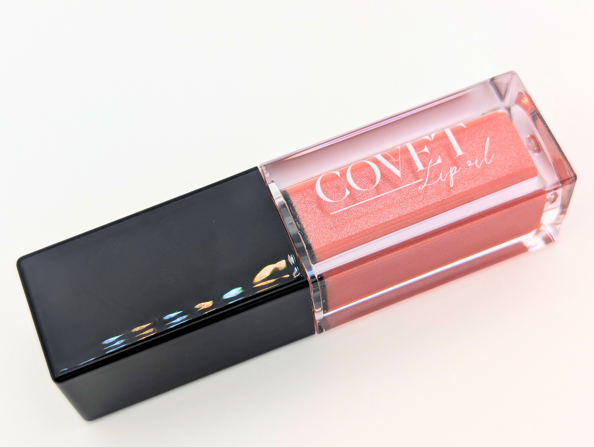 Covet Nude Pink lip gloss angled on a white background, showcasing a warm neutral pink gloss in a sleek clear tube with black cap.