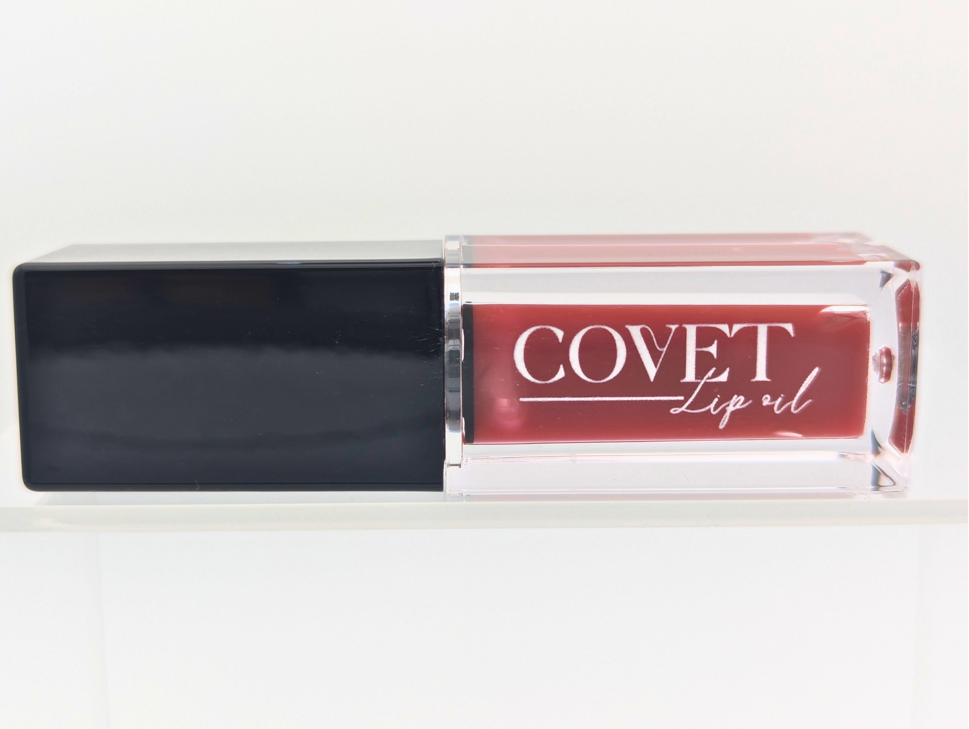 Covet Beauty’s Rosewood lip gloss — a deep rose-mauve shade with neutral brown undertones, shown in a clear gloss tube with a black cap, displayed upright on a white shelf.