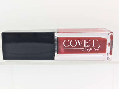 Covet Beauty’s Rosewood lip gloss — a deep rose-mauve shade with neutral brown undertones, shown in a clear gloss tube with a black cap, displayed upright on a white shelf.