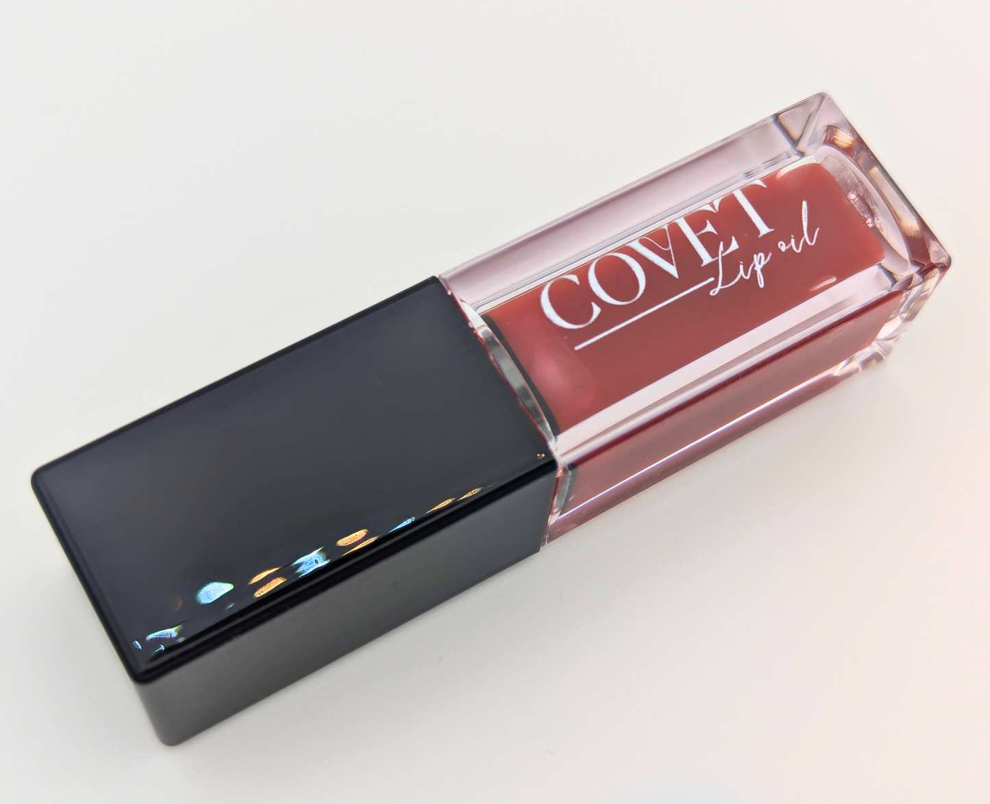 Angled view of Covet Beauty’s Rosewood lip gloss — rich mauve tone with neutral brown undertones, packaged in a clear rectangular tube with black cap on a white background.